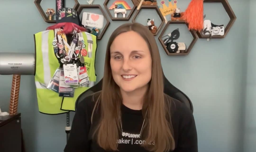 Eine Splunkerin in ihrem Büro, die ein Speaker-Shirt von der .conf23 und eine Splunk-Halskette trägt. Sie ist umgeben von Splunk-Werbeartikeln und Namensschildern von verschiedenen Splunk-Veranstaltungen.