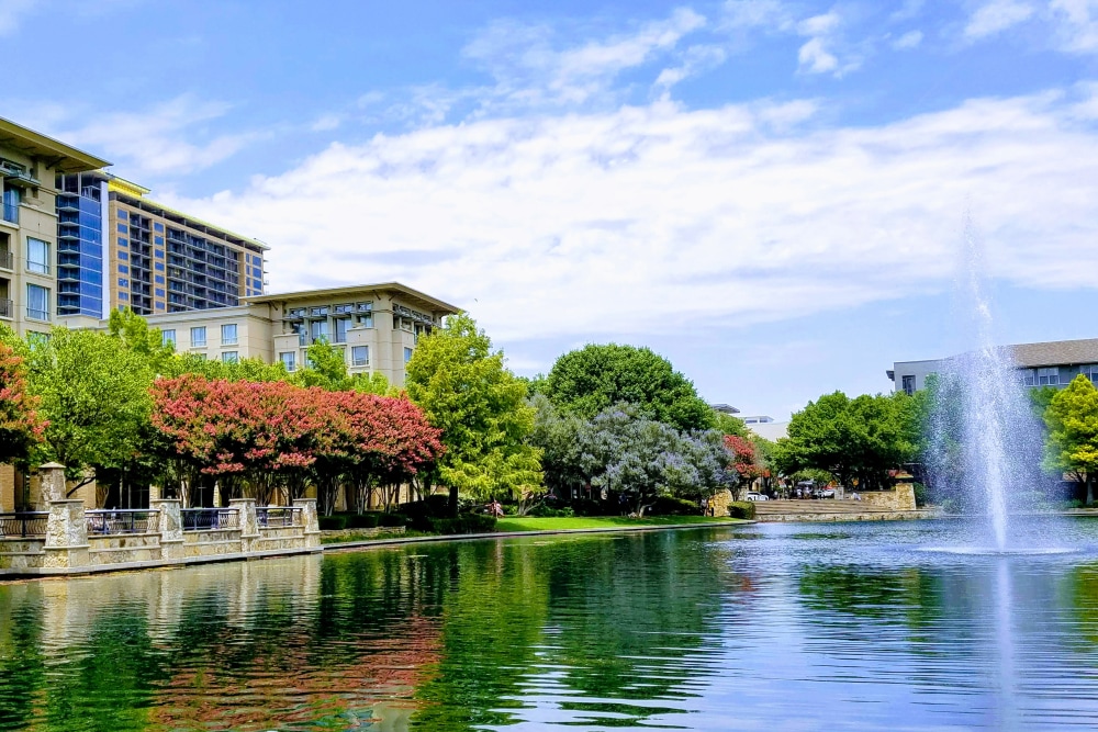  Splunk’s office in Plano, Texas. Trees and beige buildings line a large pool of water with a fountain spouting water into the air.