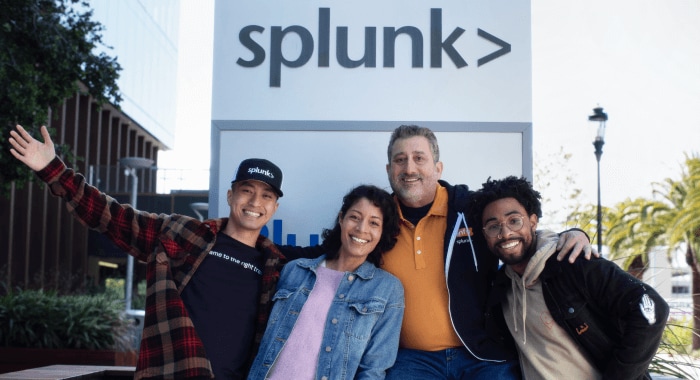 Four smiling Splunkers stand together in a celebratory stance underneath a Splunk sign.