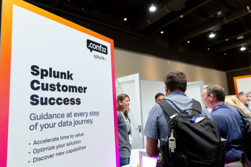  Attendees at .conf22 stopping by the Splunk customer success booth. The booth graphic has a headline that reads, “Guidance at every step of your data journey.”