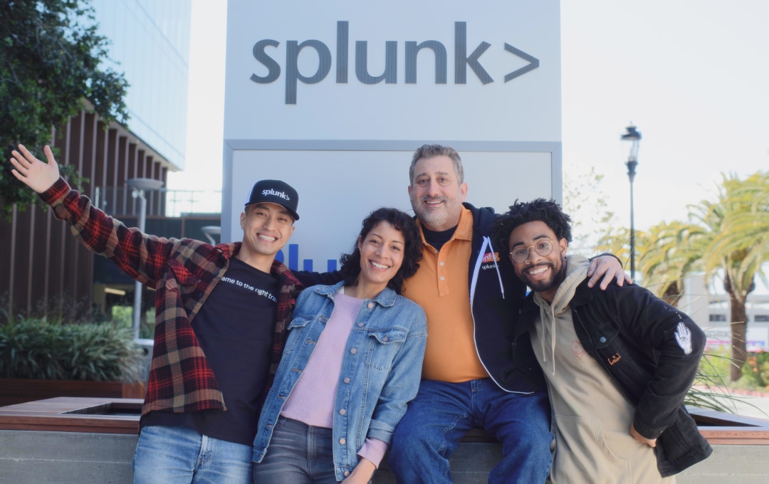  Four Splunkers smiling as they pose in front of an outdoor sign with the Splunk logo on it. 