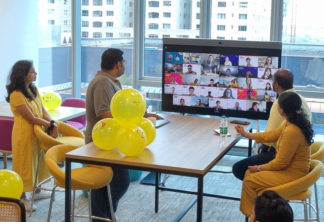 Four Splunkers surrounded by yellow balloons they’ve been using for an activity, looking towards a monitor showing a live video conference with other Splunkers.