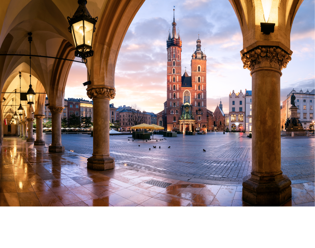 A red, two-towered church in Krakow seen through stone arches at the far end of a plaza at dusk. Lit lanterns line the arches.