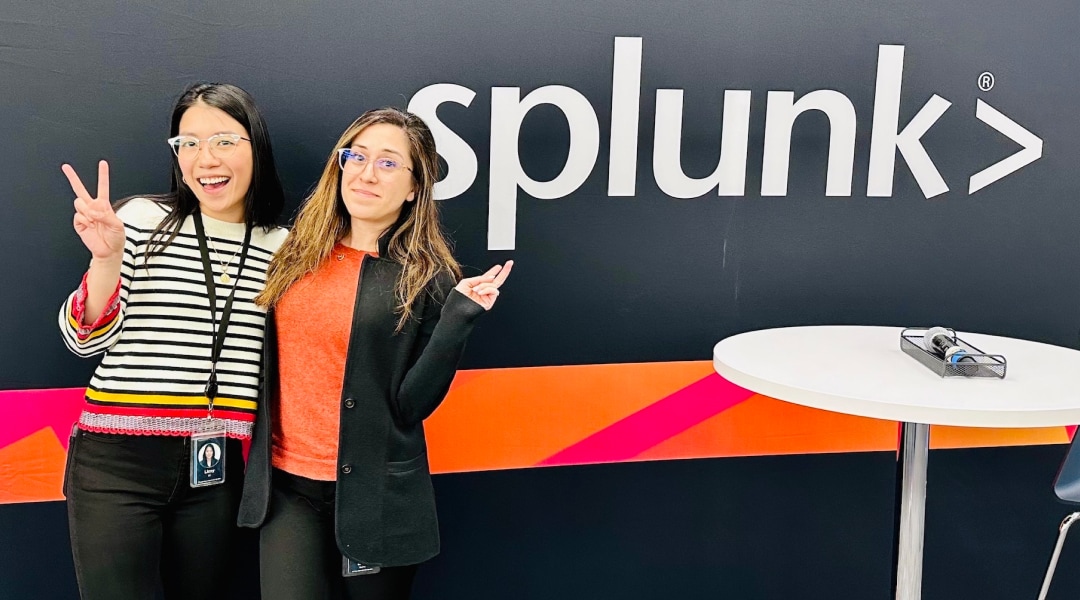 Two Splunkterns smile together as they pose in front of a wall that says Splunk. 