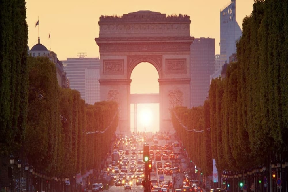 Blick durch einen großen Marmorbogen vor einer belebten sechsspurigen Straße in Paris auf die untergehende Sonne. Hohe, schlanke Bäume säumen die Straße. Splunk hat ein Büro in Paris.