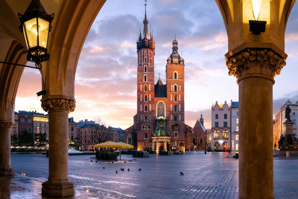 Une église rouge à deux tours est visible à travers une arche en pierre loin derrière une place au crépuscule. Des lanternes allumées sont suspendues de chaque côté de l’arche qui encadre une vue de Cracovie, en Pologne, où se trouvent des bureaux Splunk.