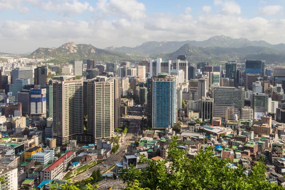 Ein Bild von Seoul, Südkorea, auf dem eine zentrale Straße eine Gruppe blauer und grüner Hochhäuser voneinander trennt, die vor einer entfernten Bergkette angeordnet ist. Splunk hat ein Büro in Seoul.