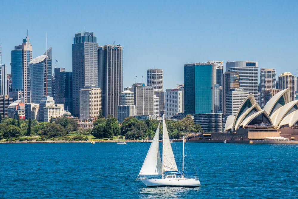 A white sailboat sails in front of a commercial area next to the Sydney Opera House. Splunk has an office in Sydney, Australia. 