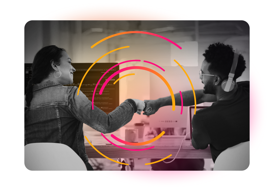 Two people, a woman and a man, are smiling and making a fist bump gesture across a desk, with computer monitors visible in the background.
