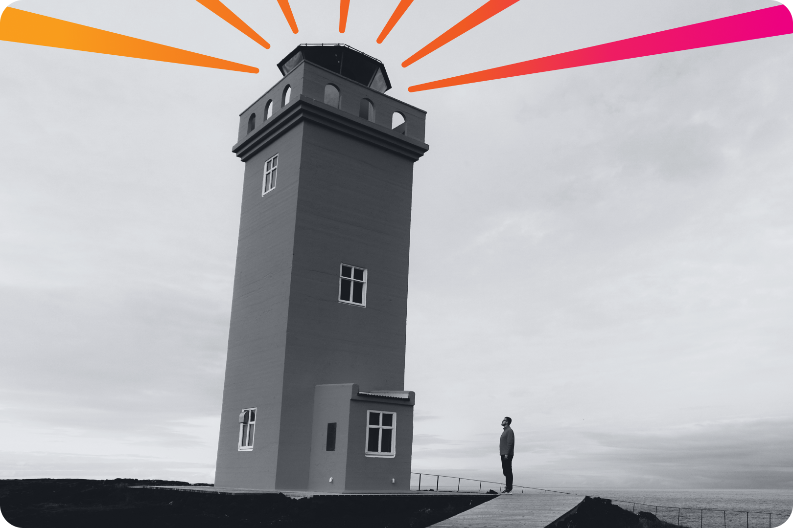 A man stands next to a tall light house which is emiting streams of gradient light from the top. 