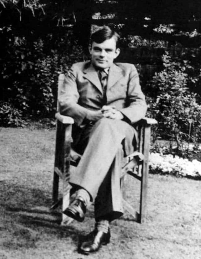 Black and white photo of a young Alan Turing with hands folded in his lap and smiling.